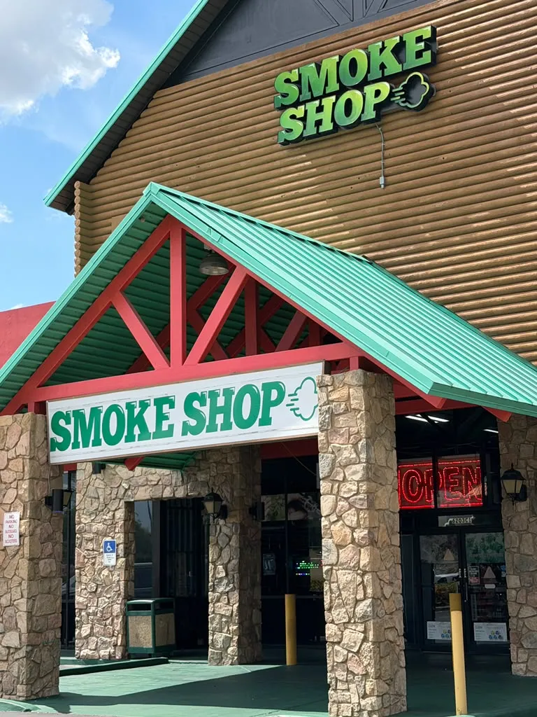 Entrance of 247 Smoke Shop with green SMOKE SHOP signs, stone columns and a lit OPEN sign on W Sand Lake Rd in Orlando