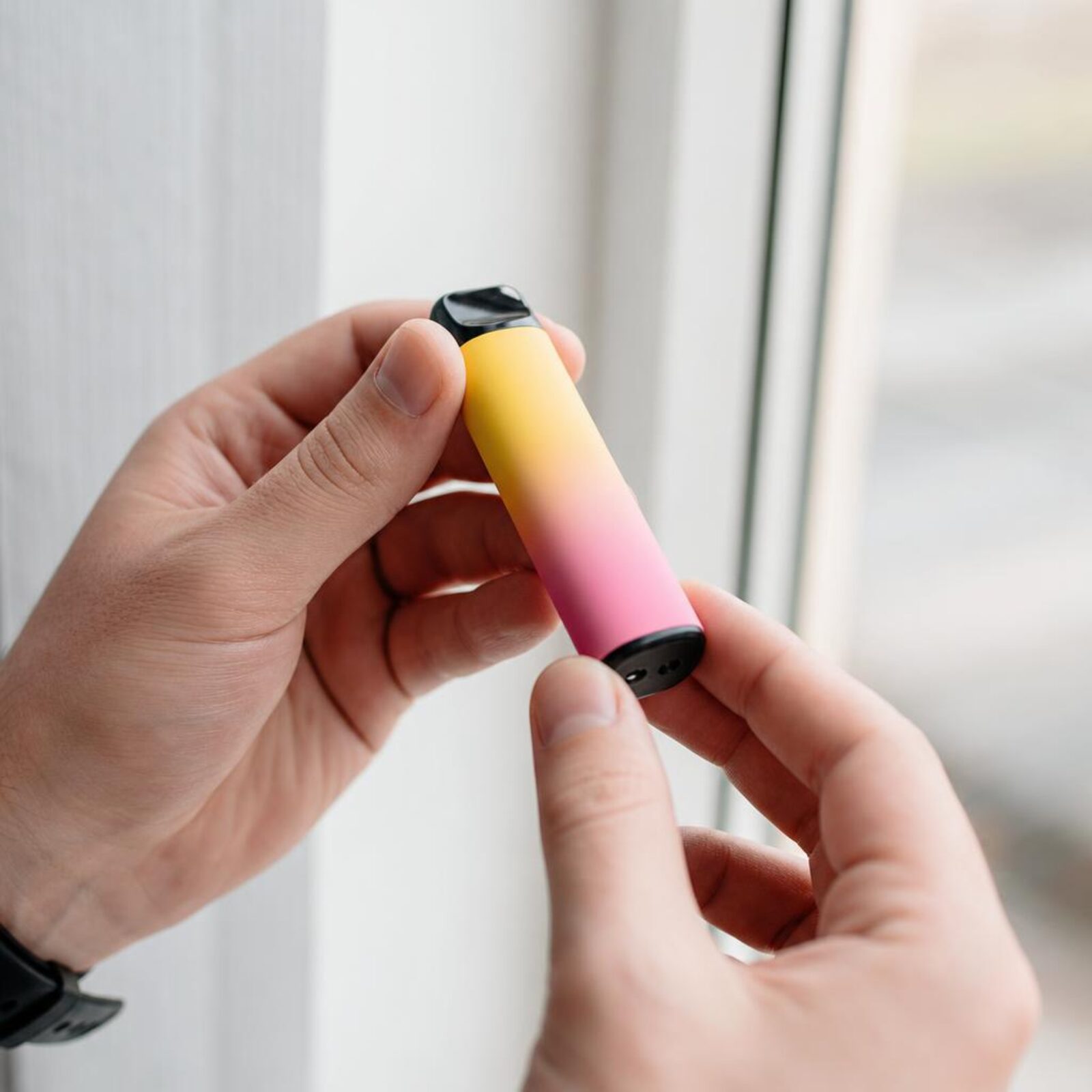Hands inspecting the mouthpiece of a disposable vape near a window.