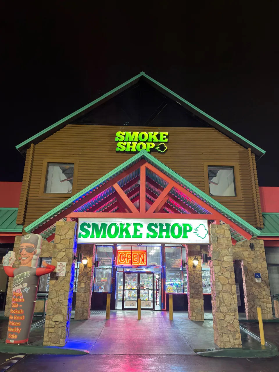 247 Smoke Shop entrance on West Sand Lake Road at night, with illuminated green signs and stone columns