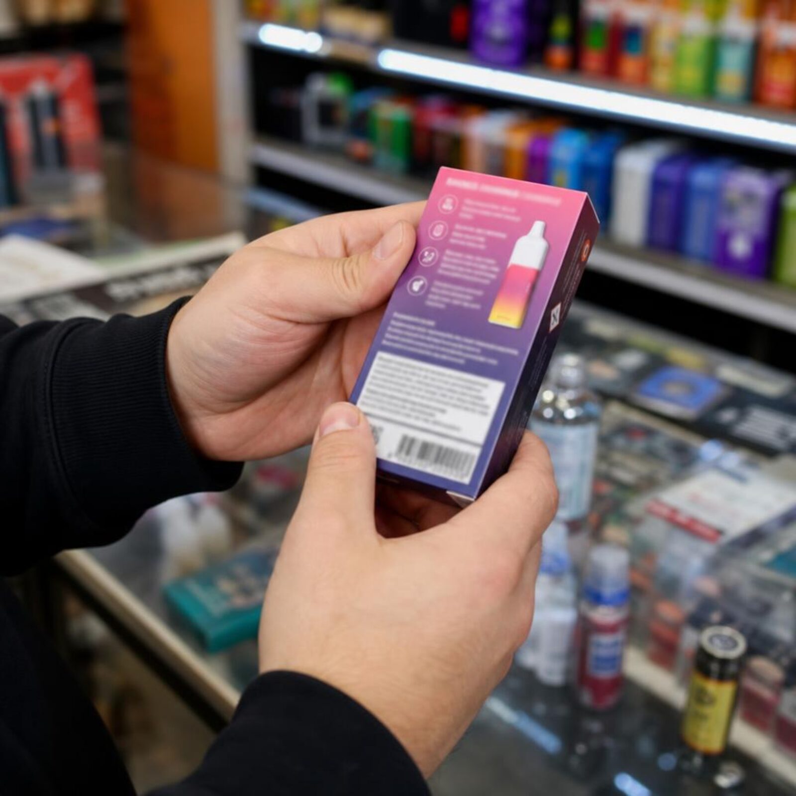 Adult hands examining the back label of a vape device box at a shop counter.